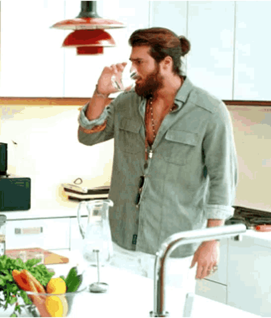 a man with a beard is drinking water from a glass in a kitchen .
