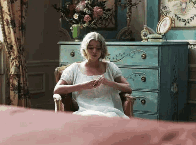 a woman in a white dress is sitting in a chair in front of a dresser in a bedroom .