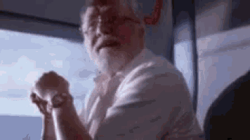 a man with a beard and glasses is sitting on a bus with his hands folded in prayer .