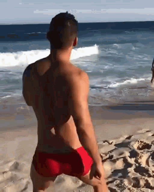a shirtless man in red swim trunks is standing on a beach .