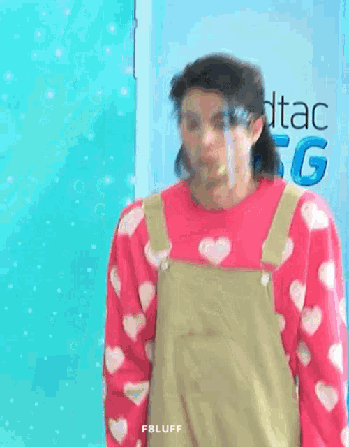 a man wearing a pink sweater with hearts on it and overalls is standing in front of a blue wall .