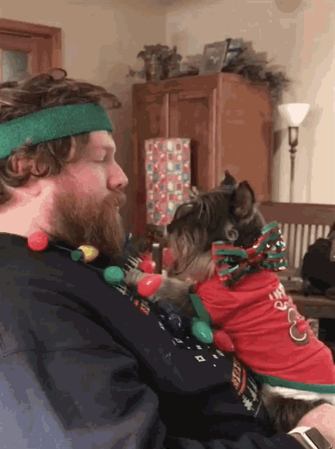 a man wearing a green headband holds a small dog wearing a sweater that says christmas