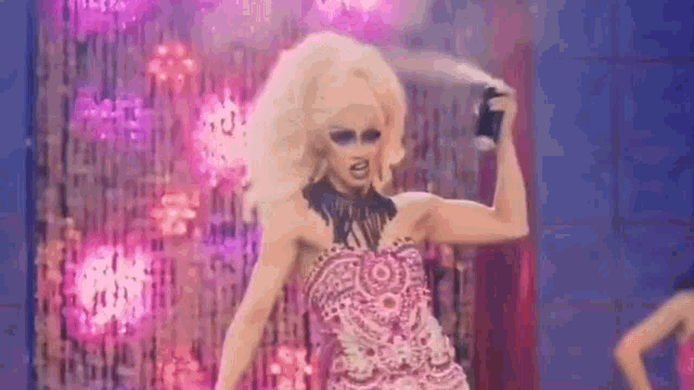 a drag queen is spraying her hair on a stage while wearing a pink dress .