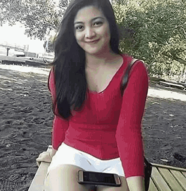 a woman in a red sweater and white shorts is sitting on a wooden bench holding a cell phone .