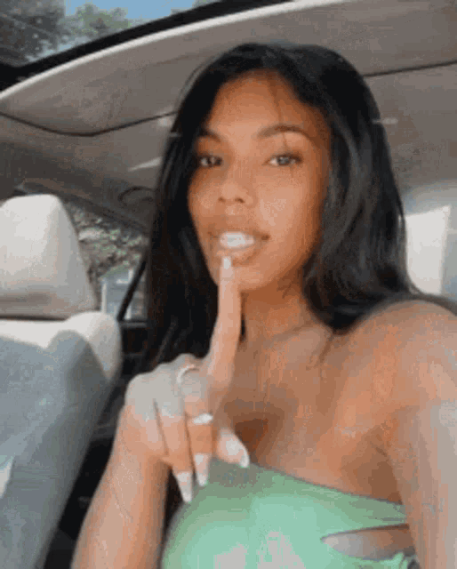 a woman in a green top is sitting in a car and holding her finger to her lips .