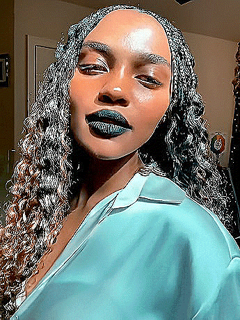 a woman with braids and blue lipstick is wearing a blue shirt and looking at the camera .