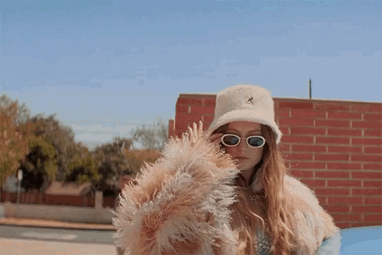 a woman wearing a kangol hat and sunglasses stands in front of a brick wall