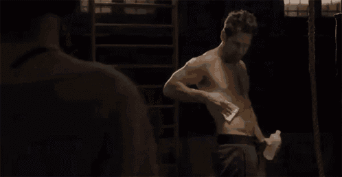 a shirtless man is standing in a dark room holding a water bottle and a towel .