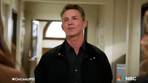 a man in a black jacket stands in a hallway with a nbc logo on the bottom