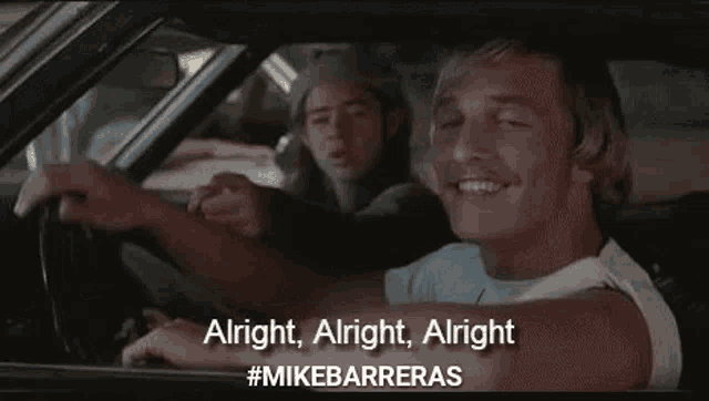a man is driving a car and pointing at the camera while another man sits behind him .