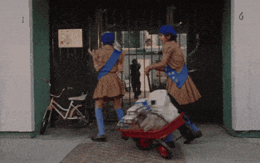 two girl scouts push a wheelbarrow full of supplies in front of a building with the number 6 on it