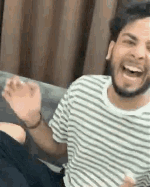 a man in a striped shirt is sitting on a couch laughing and waving .