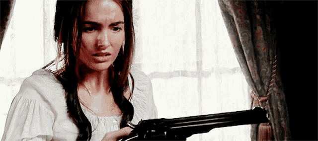 a woman in a white top is holding a gun in front of a window .