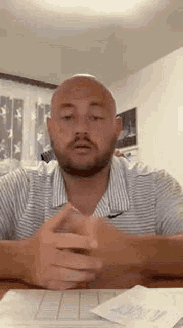 a bald man with a beard is sitting at a table with his hands folded and talking .