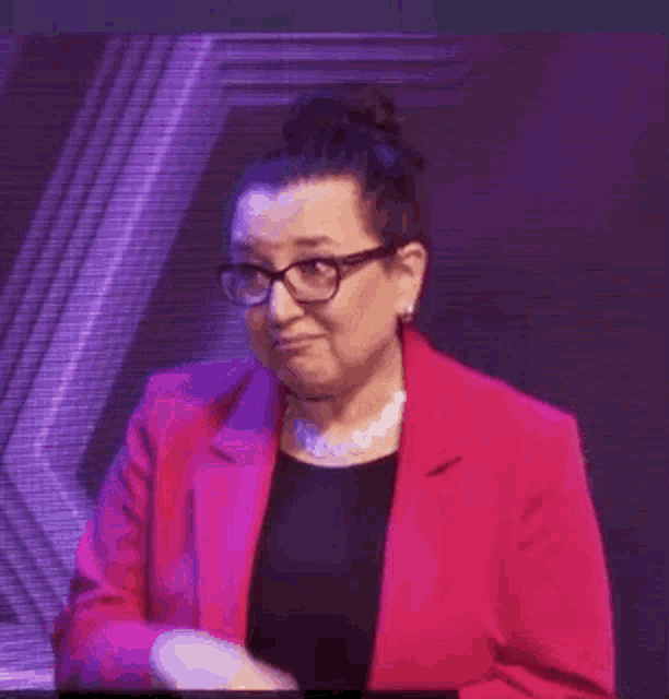 a woman wearing glasses and a pink jacket is waving her hand on a stage .