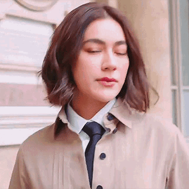 a woman wearing a trench coat and tie is standing in front of a building with her eyes closed .