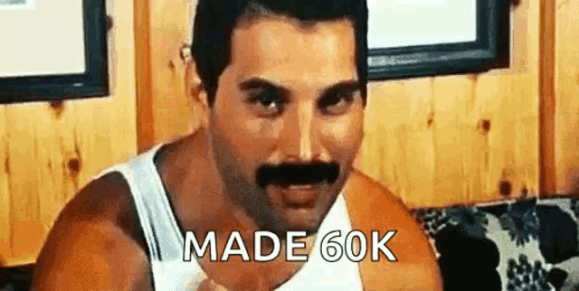 a man with a mustache wearing a white tank top is sitting on a couch and says `` made 60k '' .