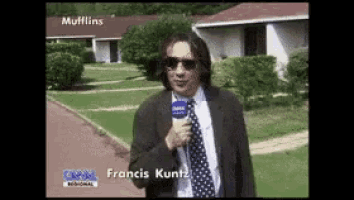 a man in a suit and tie is holding a microphone in front of a house that says " muffins "