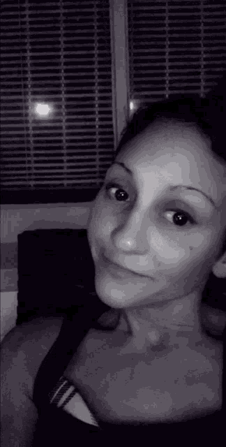 a black and white photo of a woman taking a selfie in front of a window with blinds .