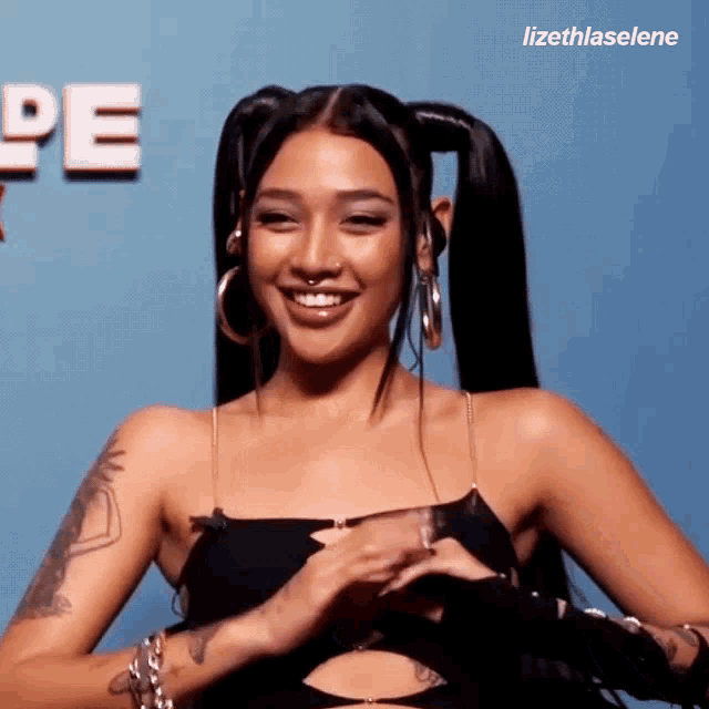 a woman with pigtails and hoop earrings is smiling in front of a blue background with the word pe on it