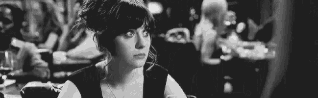 a black and white photo of a woman sitting at a table in a restaurant looking at the camera .