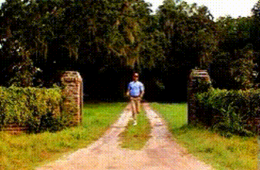 a man in a blue shirt is walking down a dirt road in front of a sign that says " welcome "