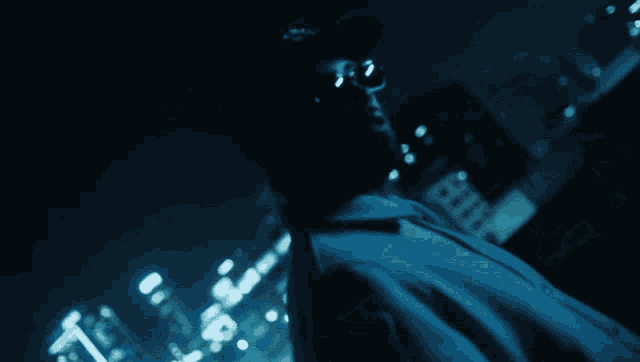 a man wearing sunglasses and a hat stands in front of a city skyline at night