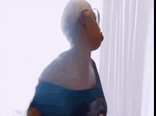 a woman wearing glasses and a blue shirt is standing in front of a white curtain .