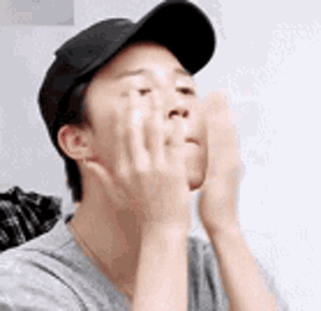 a man wearing a black baseball cap is covering his face with his hands .