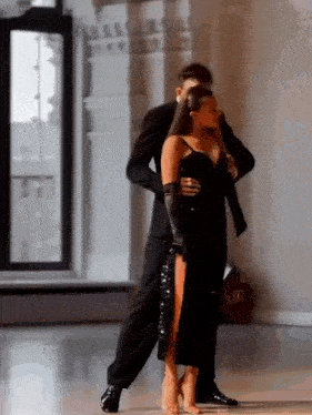 a man and a woman are dancing tango in a room . the woman is wearing a black dress with a high slit .