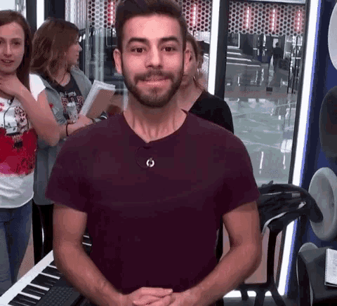 a man with a beard wearing a purple shirt is standing in front of a piano and smiling .