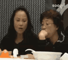 two women are sitting at a table with bowls of food and a sign that says soogif
