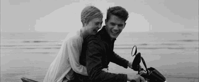 a black and white photo of a man and a woman riding a motorcycle on the beach .