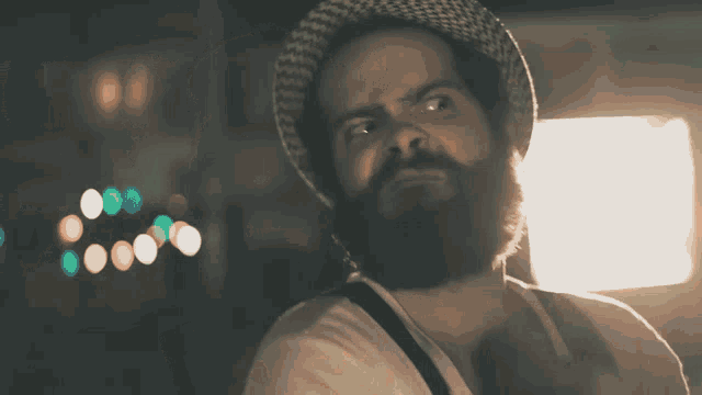 a man with a beard wearing a hat and suspenders looks at the camera