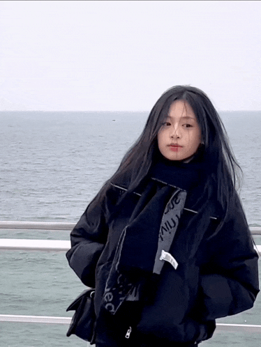 a girl wearing a black jacket and scarf stands in front of the ocean