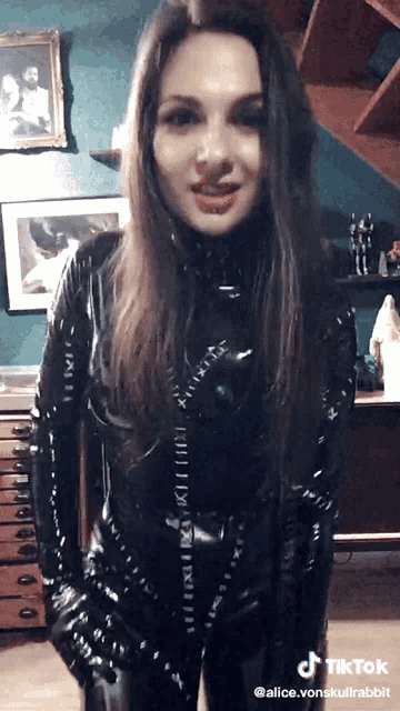 a woman in a black latex catsuit is standing in front of a staircase and has tiktok written on her face