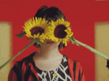 a woman in a black and white dress holds two sunflowers in front of a red wall
