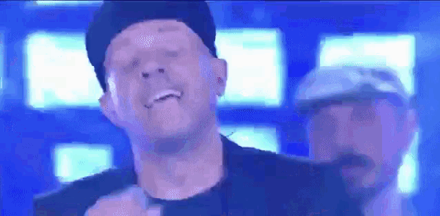 a man wearing a hat is singing into a microphone while another man stands behind him .