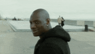 a man in a black hoodie smiles while walking down a sidewalk