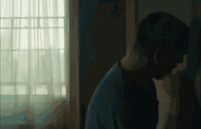 a man in a blue shirt is standing in front of a window with a white curtain .