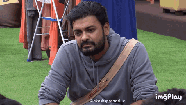 a man with a beard wearing a grey hoodie with a tan strap that says akash