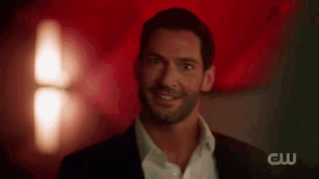 a man in a suit and white shirt is smiling in front of a red background with the cw logo in the corner .