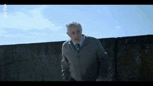 a man in a grey jacket and tie stands in front of a concrete wall with a blue sky behind him