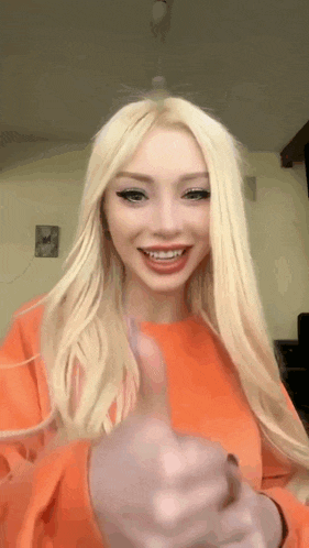 a woman with blonde hair is wearing an orange sweater and giving a thumbs up