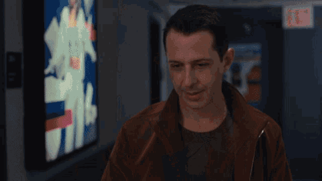a man in a brown jacket stands in a hallway with an exit sign in the background