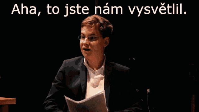 a man in a suit holds a piece of paper in front of a black background with the words aha to jste nam vysvetlil