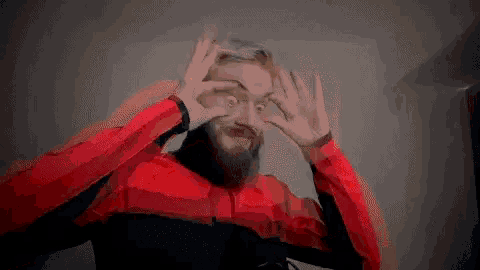 a man with a beard is wearing a red and black jacket and making a face with his hands .