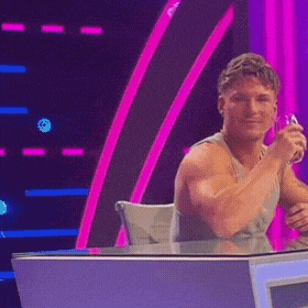 a shirtless man is sitting at a desk in front of a pink and purple backdrop