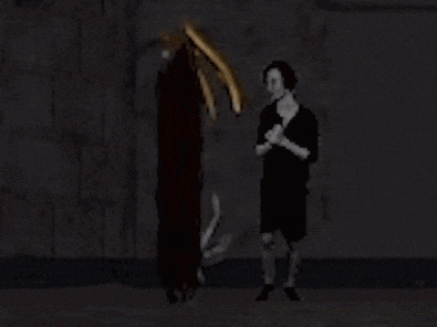 a woman in a black dress is standing in front of a skeleton in a dark room holding a sword .