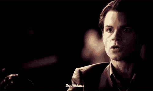 a man in a suit is talking to another man in a dark room and the man is saying `` so niklaus '' .